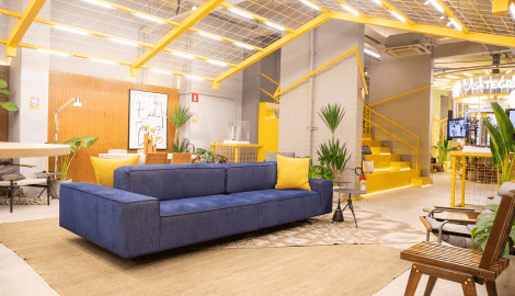 Espaço casa Tegra com um sofá azul e almofadas amarelas e tapete marron. Ao fundo do ambiente temos uma escada amarela e um painel alaranjado